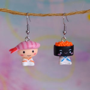 May include: Novelty earrings with sushi designs. One earring shows a shrimp character with a pink hat, the other a sushi roll with orange roe. Both have silver-coloured ear wires.