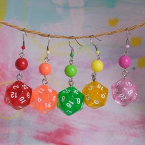Peut inclure: Cinq boucles d'oreilles en forme de dés D20 colorés suspendues à une ficelle. Chaque boucle d'oreille présente un dé de couleur différente : rouge, orange, vert, jaune et rose. Les dés ont des chiffres blancs.
