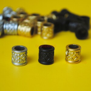 May include: Three metal beads with a decorative pattern. The beads are silver, black, and gold. The beads are on a yellow surface.