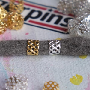 May include: Two metal hair beads, one gold and one silver, with a decorative, interwoven pattern. The beads are on a grey dreadlock.
