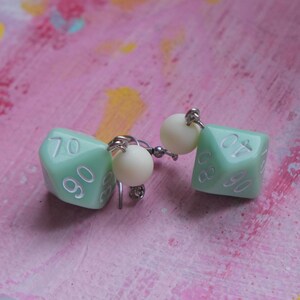 May include: A pair of dangle earrings featuring a green, six-sided dice charm with white numbers. Each earring has a white pearl bead above the dice charm.