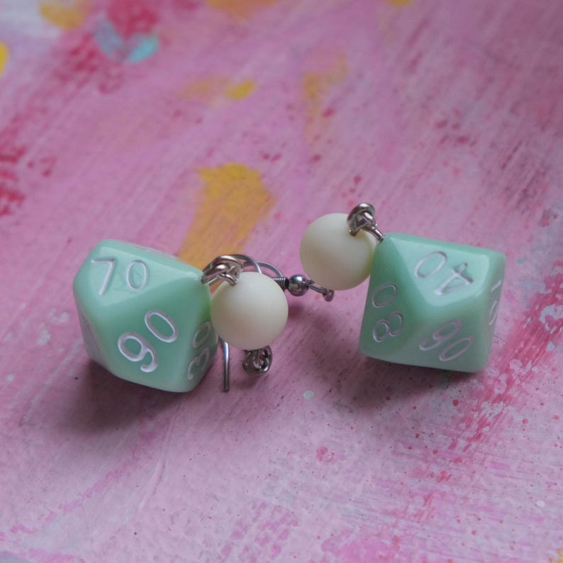 May include: A pair of dangle earrings featuring a green, six-sided dice charm with white numbers. Each earring has a white pearl bead above the dice charm.