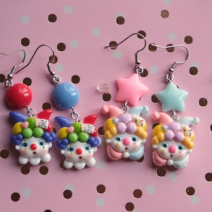May include: Four pairs of dangle earrings with a clown theme. Each earring features a clown face with colourful hair and a red nose. Some earrings have red and blue beads, while others have pink and blue star-shaped charms. The earrings hang from silver hooks.