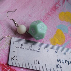 May include: A pair of earrings featuring a green, 20-sided die charm and a white bead. The earrings are hanging from silver hooks.