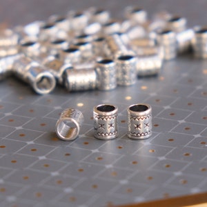 May include: Silver-colored metal tube beads with etched patterns. The beads have a cylindrical shape with a hole running through the center. Some beads have a star and cross design. A collection of beads are scattered on a gray surface.
