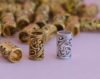 May include: Close-up of decorative metal beads in gold and silver tones. The beads have an open cylinder shape with intricate scrollwork designs. A few beads are in focus, while others are blurred in the background, suggesting a craft supply.