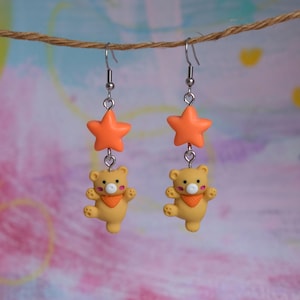 May include: A pair of dangle earrings featuring orange star charms and yellow bear charms with orange neckerchiefs. The earrings are suspended from silver-colored hooks, creating a playful and whimsical accessory.