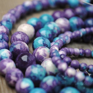 May include: A close-up of a strand of purple and blue gemstone beads. The beads are round and have a marbled pattern.
