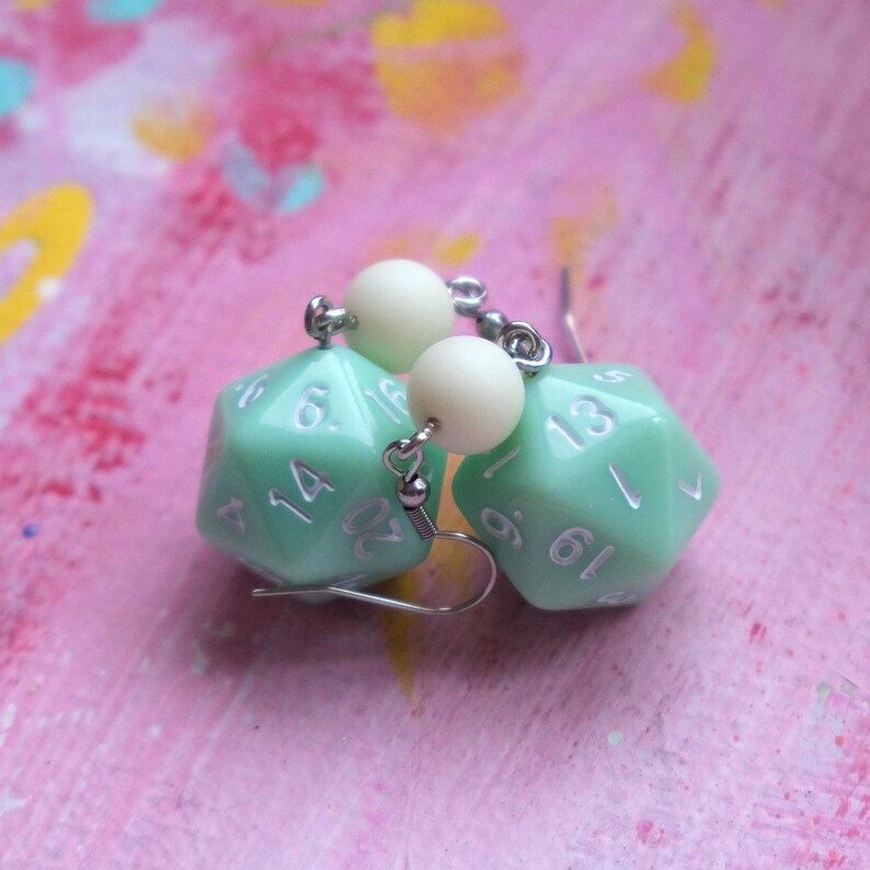 May include: A pair of earrings featuring a light green 20-sided dice charm with white numbers and a white bead on each earring.