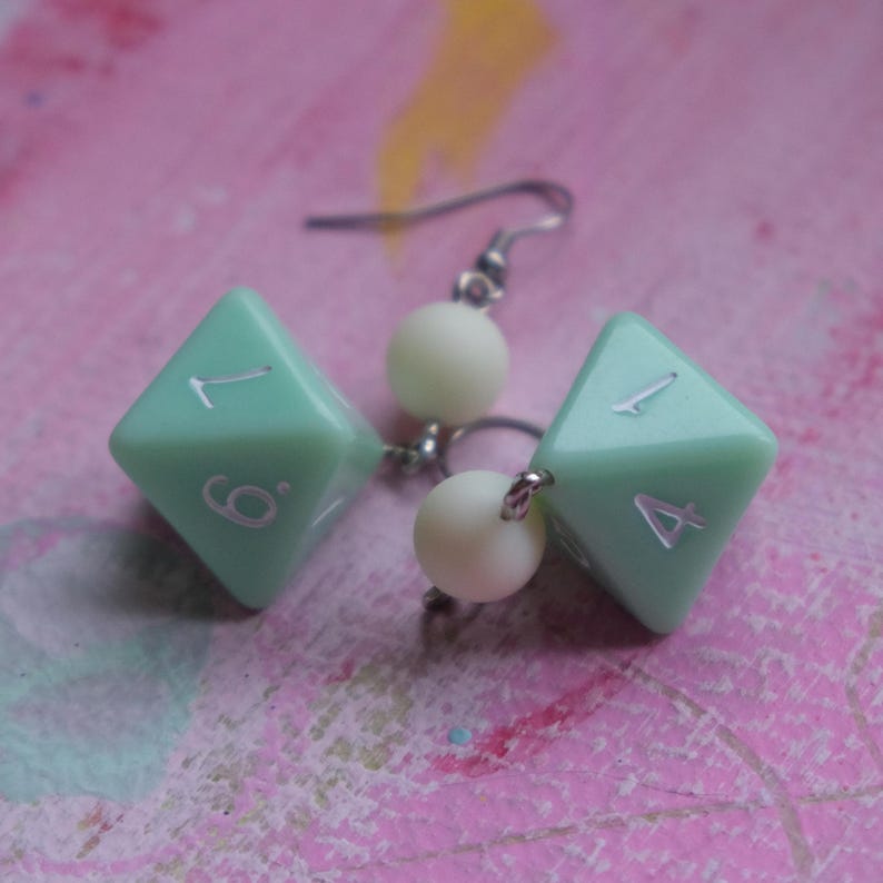 May include: A pair of earrings featuring a light green dice charm with white numbers on each side. The dice are attached to a silver hook with a white pearl bead.