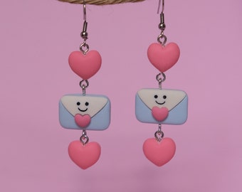 May include: A pair of dangling earrings featuring a pink heart, a light blue envelope with a smiling face, and another pink heart. The earrings are suspended from a silver-colored hook and are set against a pink background.