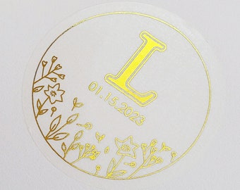 May include: A white round sticker with a gold floral design and the letter "L" in the center. The date "01.15.2023" is printed below the letter.