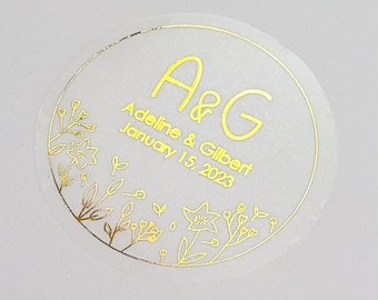 May include: A clear round sticker with gold lettering and floral designs. The sticker says "A&G, Adeline & Gilbert, January 15, 2023".