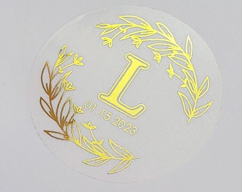 May include: A clear round sticker with a gold floral wreath design and the letter "L" in the center. The date "01.15.2023" is printed below the letter.