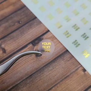 May include: A clear, round sticker with gold foil text that reads "YOUR TEXT HERE." The sticker is held by silver tweezers. A sheet of similar stickers is in the background, along with a wooden surface.