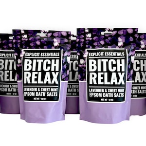 Op de afbeelding: Meerdere zakjes Explicit Essentials Bitch Relax Lavendel & Zoete Munt Epsom badzout. Elk zakje heeft een paars bloemenpatroon aan de bovenkant, een wit label met dikke zwarte tekst en een lichtpaarse onderkant, met een nettogewicht van 340 gram.