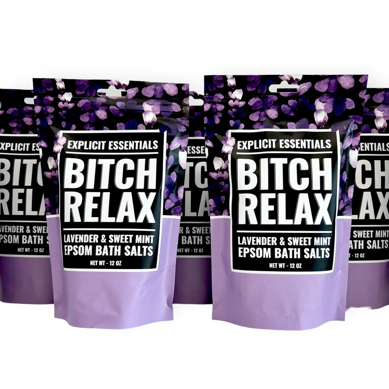 Op de afbeelding: Meerdere zakjes Explicit Essentials Bitch Relax Lavendel & Zoete Munt Epsom badzout. Elk zakje heeft een paars bloemenpatroon aan de bovenkant, een wit label met dikke zwarte tekst en een lichtpaarse onderkant, met een nettogewicht van 340 gram.