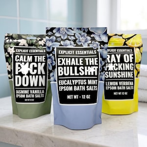 May include: Three pouches of Explicit Essentials Epsom bath salts, each 12 oz. The left is green with jasmine vanilla, the center is blue with eucalyptus mint, and the right is yellow with lemon verbena, all featuring bold text and floral patterns.