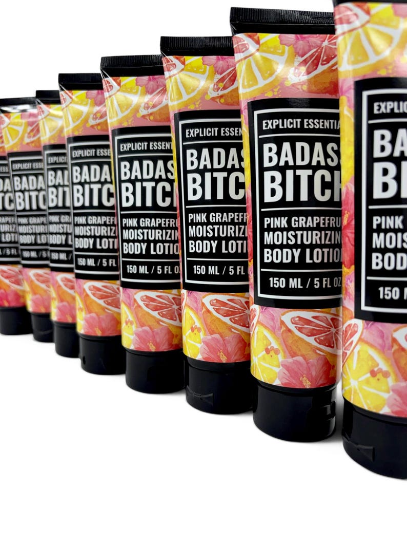 May include: A row of squeeze tubes containing Explicit Essentials Badas Bitch Pink Grapefruit Moisturizing Body Lotion. Each tube features a vibrant pattern of pink grapefruit slices and hibiscus flowers, with a black cap and 150 ml / 5 fl oz label.