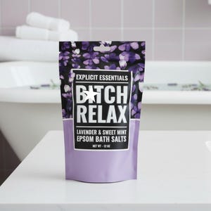 Op de afbeelding: Een stazak met Explicit Essentials Epsom-badzout in lavendel en zoete munt. De verpakking heeft een paars bloemenpatroon, een wit label met de zwarte tekst BITCH RELAX en een nettogewicht van 12 oz dat op de voorzijde is gedrukt.