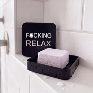 May include: A square, light purple bar of soap rests in a black, square soap dish. Behind the dish stands a black square sign with white text that reads F*CKING RELAX, featuring a white star symbol in place of the letter U in the first word.