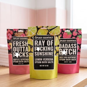 May include: Three stand-up pouches of Explicit Essentials Epsom bath salts, each 12 oz. The left is pink with tropical fruit, the center is yellow with lemon graphics, and the right is pink with raspberries. Each label features bold text.