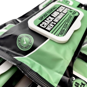 May include: A stack of black and green packs of intimate wipes. The packaging features the text "CRACK AND SACK MEN'S INTIMATE WIPES" in white and black, with a green rectangular label. The packaging also includes the text "FRESH - CLEAN - REFRESHING PACK".