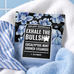 May include: A black package of Eucalyptus Mint shower steamers with blue floral designs and the text "EXHALE THE BULLSH*T". The package contains 4 tablets, each weighing 25 grams. The package is resting on a white towel and blue striped fabric.
