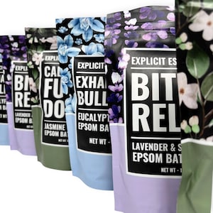 May include: A row of colorful bath salt pouches with floral designs. The pouches feature text like "Calm", "Exhale", and "Bit Rel". Each pouch has a different color scheme, including blue, green, and purple, with a black top.