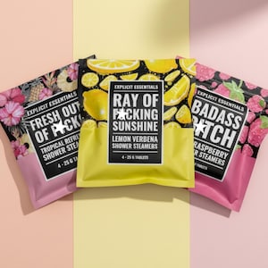 May include: Three rectangular pouches of shower steamers labeled Explicit Essentials. The left pouch is pink and floral, the center is yellow with lemon graphics, and the right is pink with raspberry graphics. Each contains 4-25 g tablets.