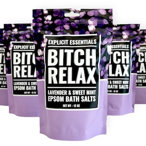 Op de afbeelding: Meerdere zakjes Explicit Essentials Bitch Relax Lavendel & Zoete Munt Epsom Badzout. Elk zakje heeft een zwart en paars bloemenpatroon, een wit label met dikke zwarte tekst en een nettogewicht van 340 gram per verpakking.