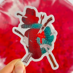 May include: A red crocheted heart with various tools and weapons piercing through it. The heart is surrounded by a white border.