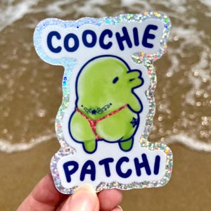 May include: A sticker with the words "COOCHIE PATCHI" in blue letters. The sticker features a green cartoon character wearing a red thong with a star design. The sticker has a holographic, glittery border.