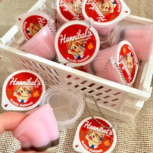 May include: A white basket filled with pink pudding cups, each with a red and white label that reads "Hannibal's Blood Pudding." The pudding has a pink top and a brown base. Some cups have a silver chain attached.