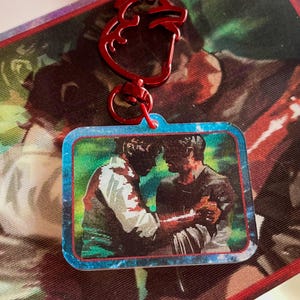 May include: A red heart-shaped keychain with a rectangular charm featuring a pixelated image of two people. The charm has a blue border and is attached to a patterned background.
