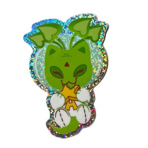 May include: A green cat-like character pin with a star-shaped object. The pin has a glittery, iridescent border and features a cat-like face with a red triangle on its forehead. The cat is holding a yellow star with the letter "A" on it.