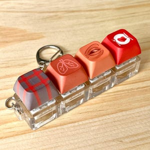 May include: A keychain featuring four custom keyboard keycaps. The keycaps are in shades of red, orange, and gray, with various designs including a plaid pattern, lungs, lips, and a plate with a knife and fork.