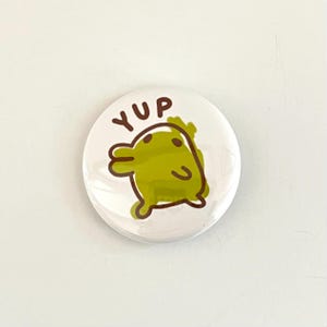 May include: White button with a cartoon character saying "YUP" in brown text. The character is green and has a brown outline.