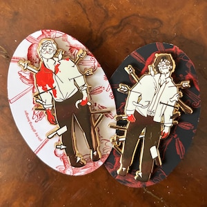 Może przedstawiać: Dwie emaliowane przypinki przedstawiające postać przebitą strzałami i nożami. Jedna przypinka ma czerwono-białe tło, druga czarno-czerwone. Postacie noszą białe koszule, czarne spodnie i złote akcenty.