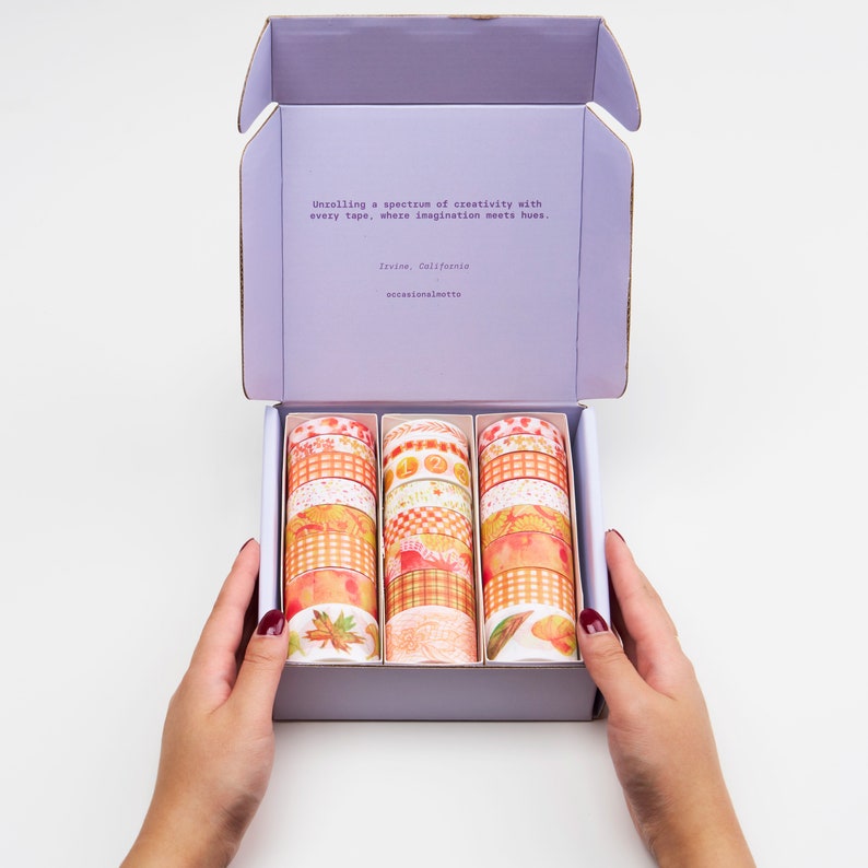 May include: A box of colourful washi tape rolls with autumnal designs, including leaves, plaid, and floral patterns. The box is open and the rolls are arranged in rows. The box is labelled with the text "Unrolling a spectrum of creativity with every tape, where imagination meets hues. Irvine, California occasionalmotto"