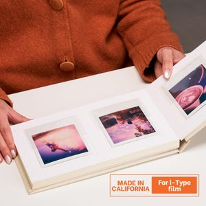 以下が含まれることがあります： 3枚のi-Typeフィルム写真が表示された、開いたクリーム色のフォトアルバム。アルバムは白い内側です。写真は様々な画像を表示しています。オレンジ色のセーターを着た人がアルバムを持っています。アルバムには「Made in California」と記載されています。