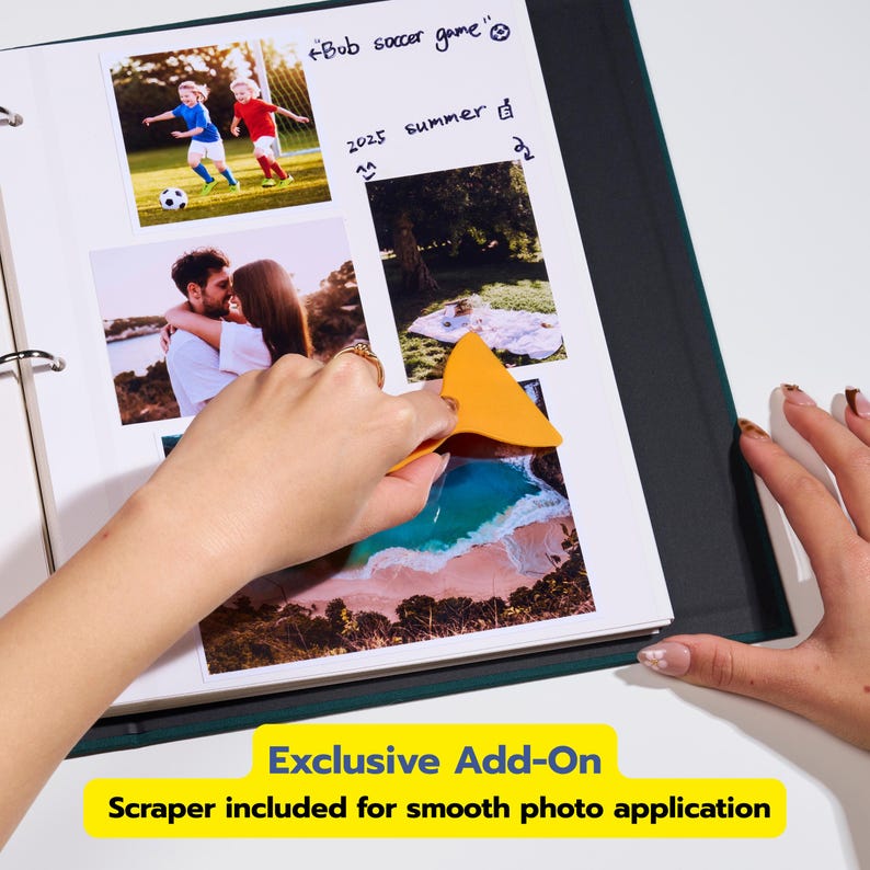May include: A dark green photo album with white pages containing various photographs and handwritten notes including "Bob soccer game" and "2025 summer". An orange plastic scraper tool is used to smooth a photo onto the page surface.