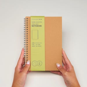May include: A brown spiral notebook with a green label that says "Occasional Mette Minimal Collection Spiral-Kraft Notebook Large (5.5 x 8.3 in) Blank 100 Pages 100gsm Eco-Friendly".