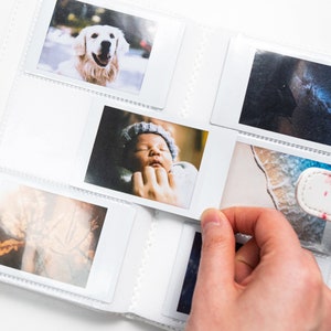 May include: A white photo album with clear plastic sleeves for holding photos. The album is open and several photos are visible, including a photo of a dog, a baby, and a beach scene.