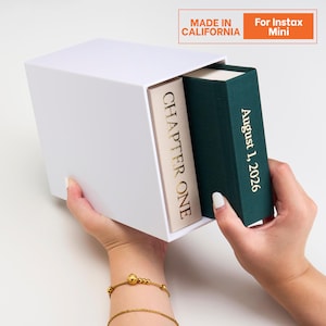 May include: A white rectangular storage box containing two book-shaped containers. One is a cream-colored spine labeled CHAPTER ONE and the other is a dark green spine with gold text reading August 1, 2026. The box is designed for Instax Mini.
