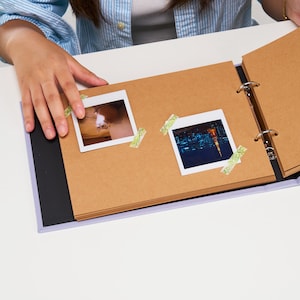 May include: A brown photo album with a black spine and a purple border. The album is open to a page with two photos, one of a cat and one of a city skyline at night. Both photos are decorated with green washi tape.