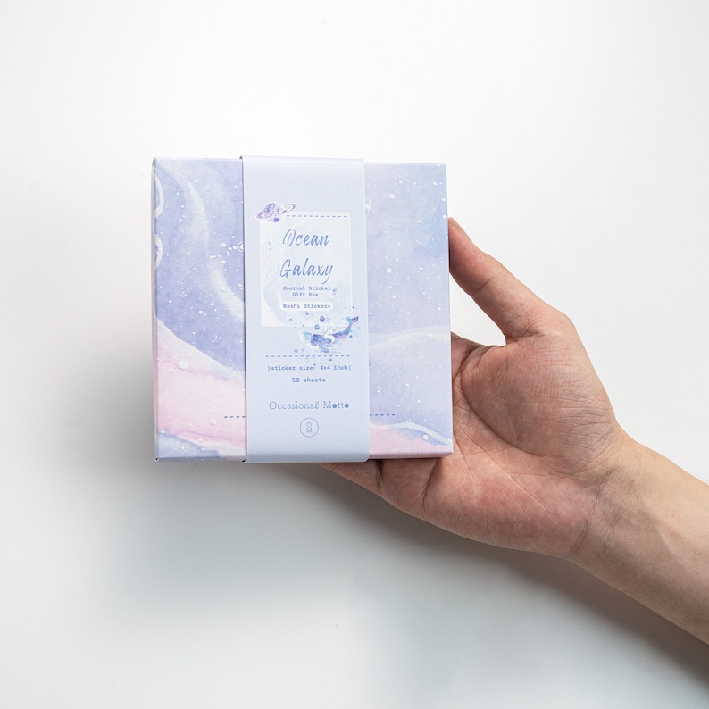 May include: A box of "Ocean Galaxy" journal stickers with a watercolor design of a galaxy in shades of blue and pink. The box has a sticker with the text "Journal Stickers, 50 sheets,  Washable Stickers, Sticker size: 1.5" x 1.5", 50 sheets, Occasional Matte"