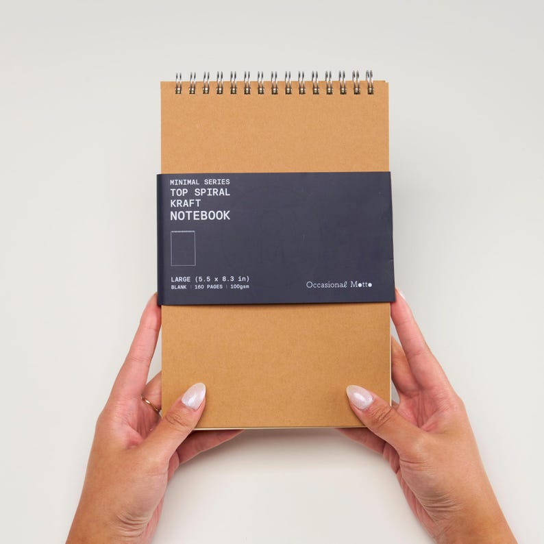 Op de afbeelding: Een bruin kraft notitieboek met een zwart-wit label met de tekst "Minimal Series Top Spiral Kraft Notebook Large (5.5 x 8.3 in) Blank - 100 Pages - 100gsm Occasional Matte"