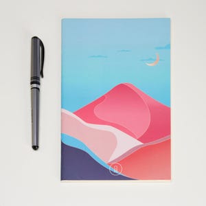 May include: A pink and blue illustrated notebook with a crescent moon and a mountain range design. The notebook has a white background and a small logo in the bottom right corner.
