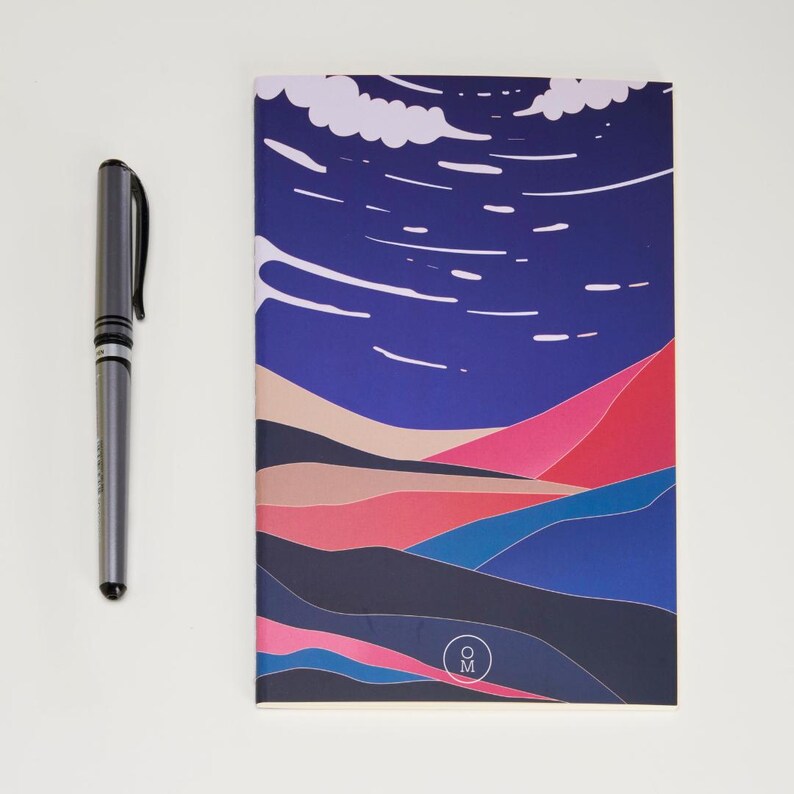 May include: A blue and pink notebook with a stylized landscape design. The cover features a dark blue sky with white clouds and a series of pink, blue, and brown hills. The notebook has a small circle with the letters "O M" in the lower right corner.
